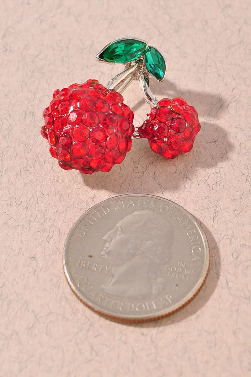 A cherry-shaped brooch covered in red rhinestones with green marquise-cut stones for leaves, set on a silver-tone metal and displayed on a textured beige surface next to a quarter for size comparison.