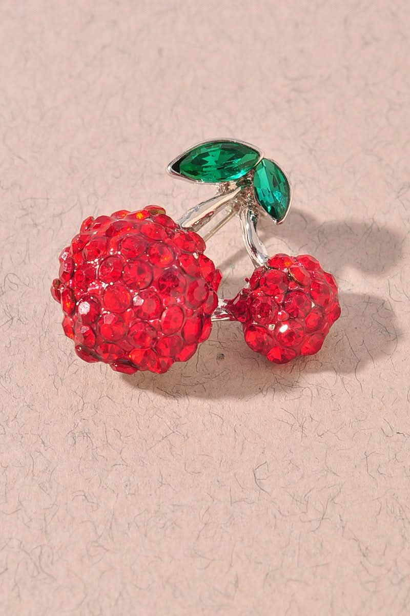 A cherry-shaped brooch covered in red rhinestones with green marquise-cut stones for leaves, set on a silver-tone metal and displayed on a textured beige surface.