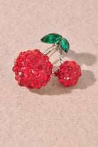 A cherry-shaped brooch covered in red rhinestones with green marquise-cut stones for leaves, set on a silver-tone metal and displayed on a textured beige surface.