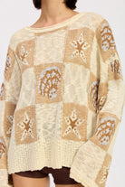 Woman wearing a cream and taupe patchwork knit sweater with star and seashell motifs, closeup front view. and brown knee-high boots on a white background