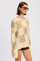 Woman wearing a cream and taupe patchwork knit sweater with star and seashell motifs, front view. and brown knee-high boots on a white background