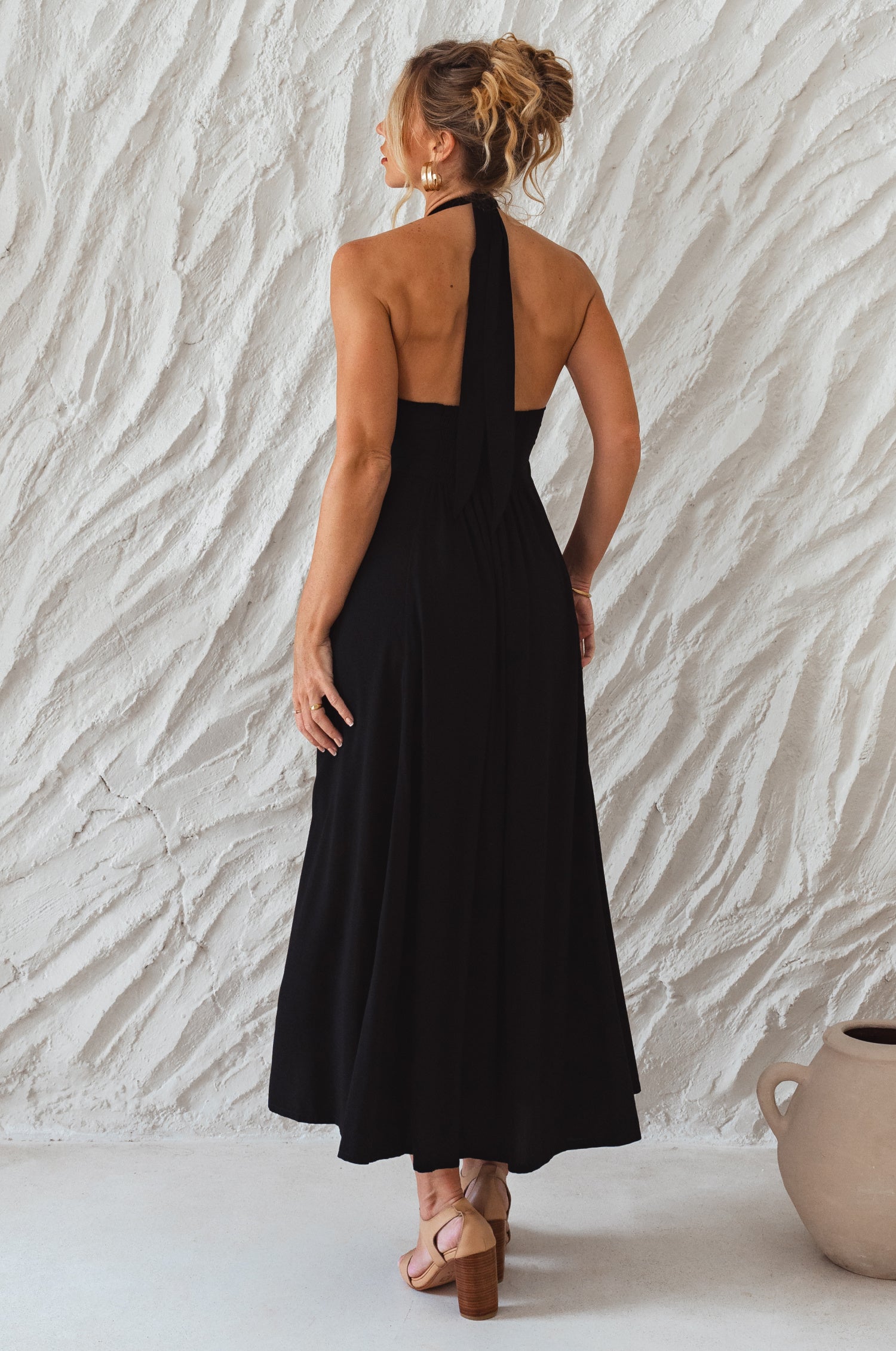 Woman wearing a black soft rayon maxi dress with halter neckline, smocked back, and fitted bodice, on a white background, back view