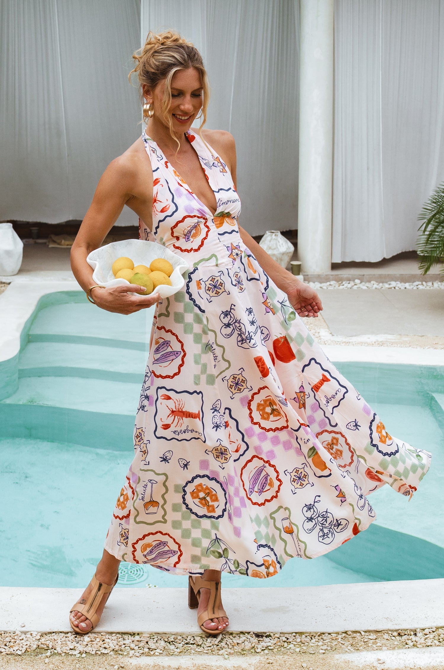 Woman wearing a white soft rayon whimsical patterned maxi dress with halter neckline, smocked back, and fitted bodice, by a pool, front view