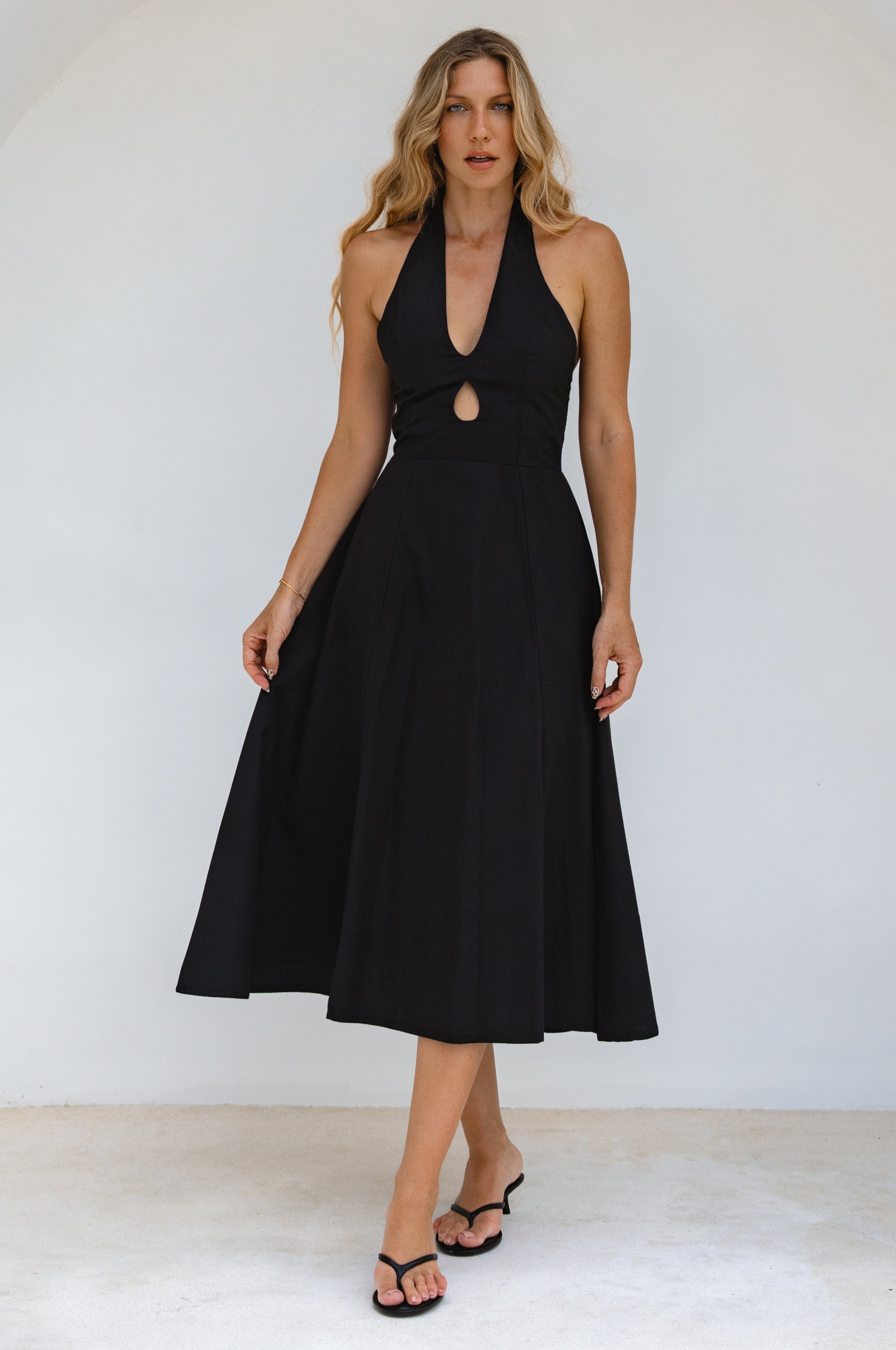 Woman wearing a sleeveless black cotton midi dress with halter neckline against a white background