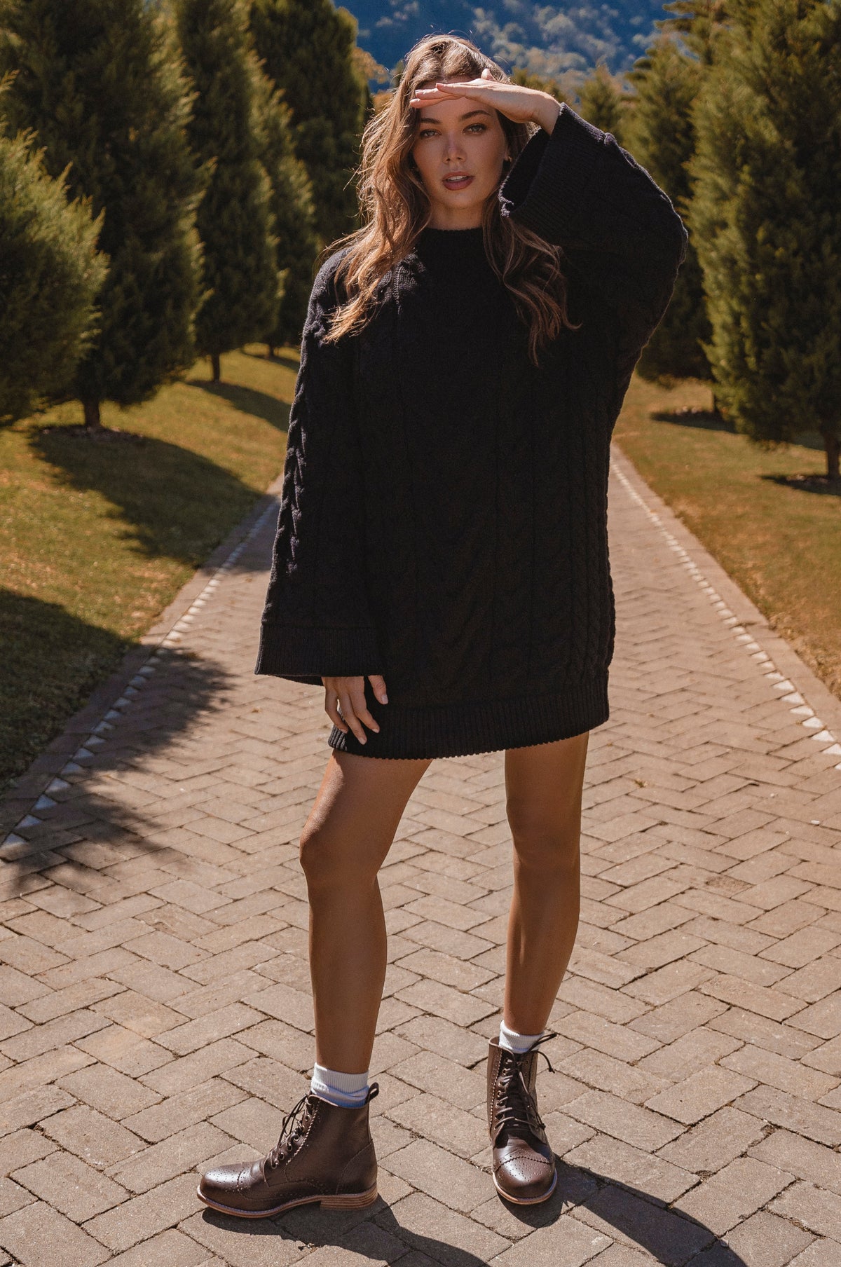 Woman in a black cable knit sweater dress standing on a paved path with trees in the background