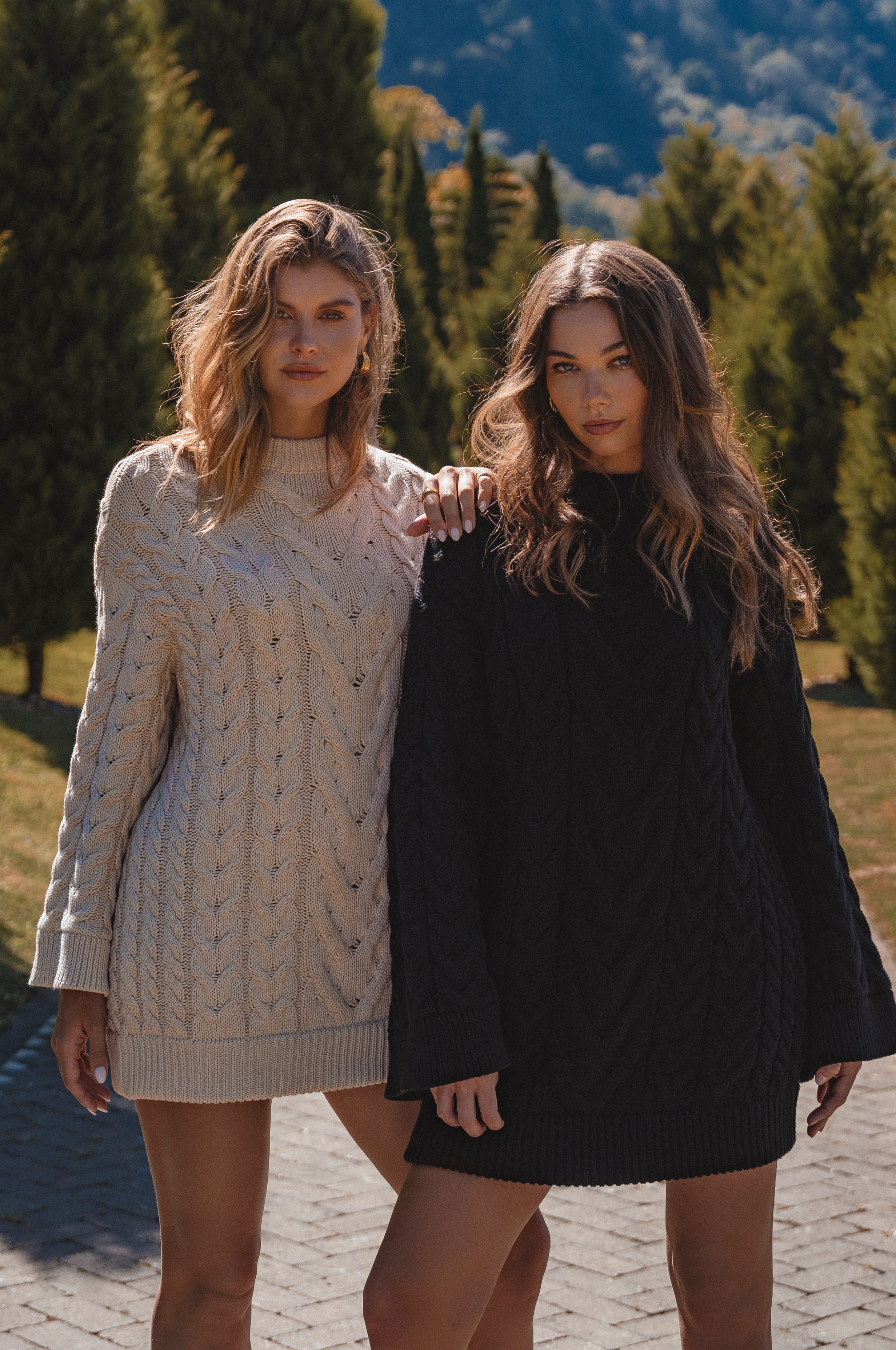 Two women standing outdoors with trees and mountains in the background wearing cable knit sweater dresses