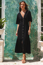 Woman wearing black midi dress in a button-down silhouette with a smocked back and fitted front, tiered skirt, and butterfly sleeves standing in front of a green wall, front view