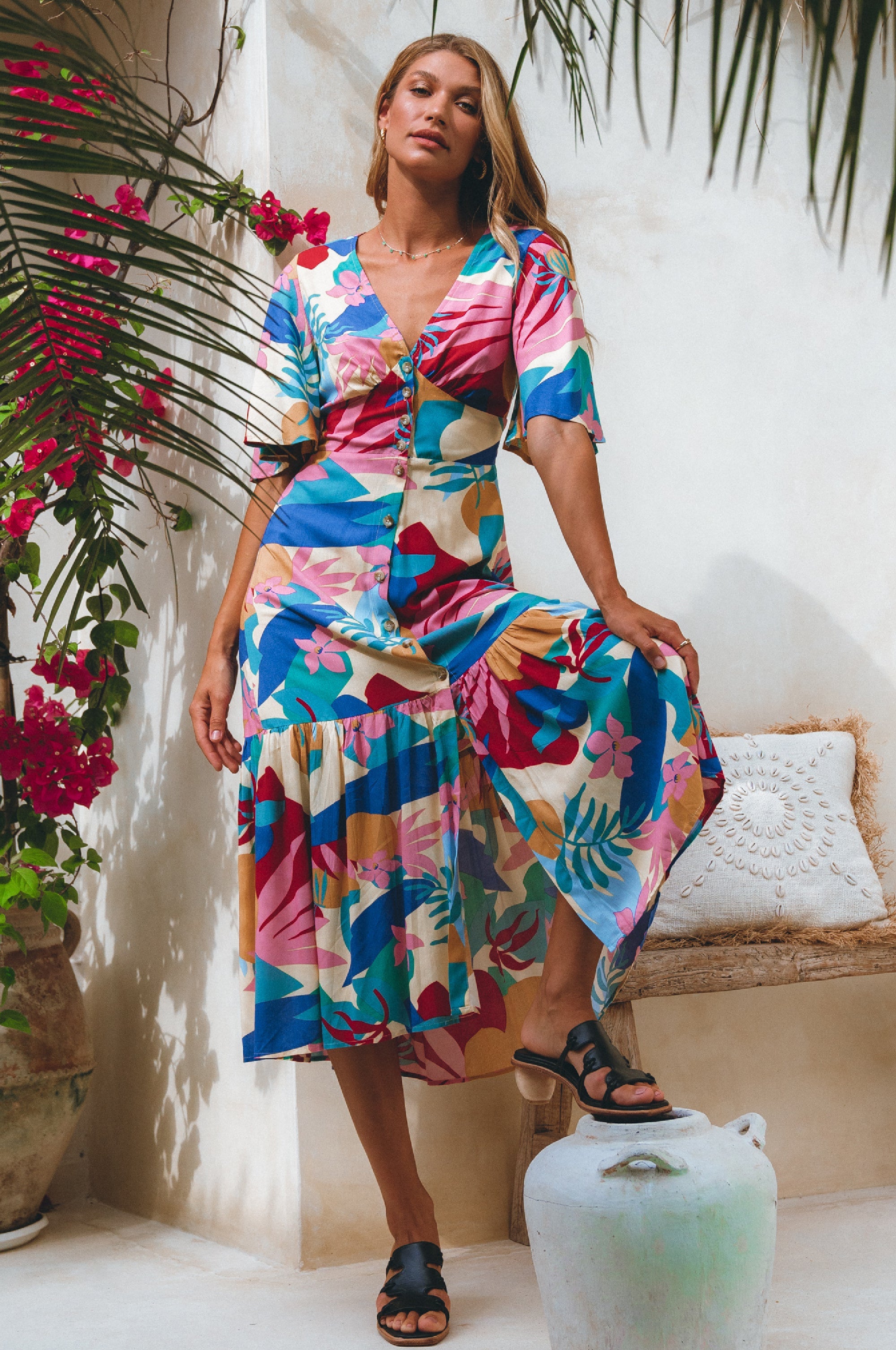 Woman wearing blue, pink and yellow patterned midi dress in a button-down silhouette with a smocked back and fitted front, tiered skirt, and butterfly sleeves standing against a neutral background, front view