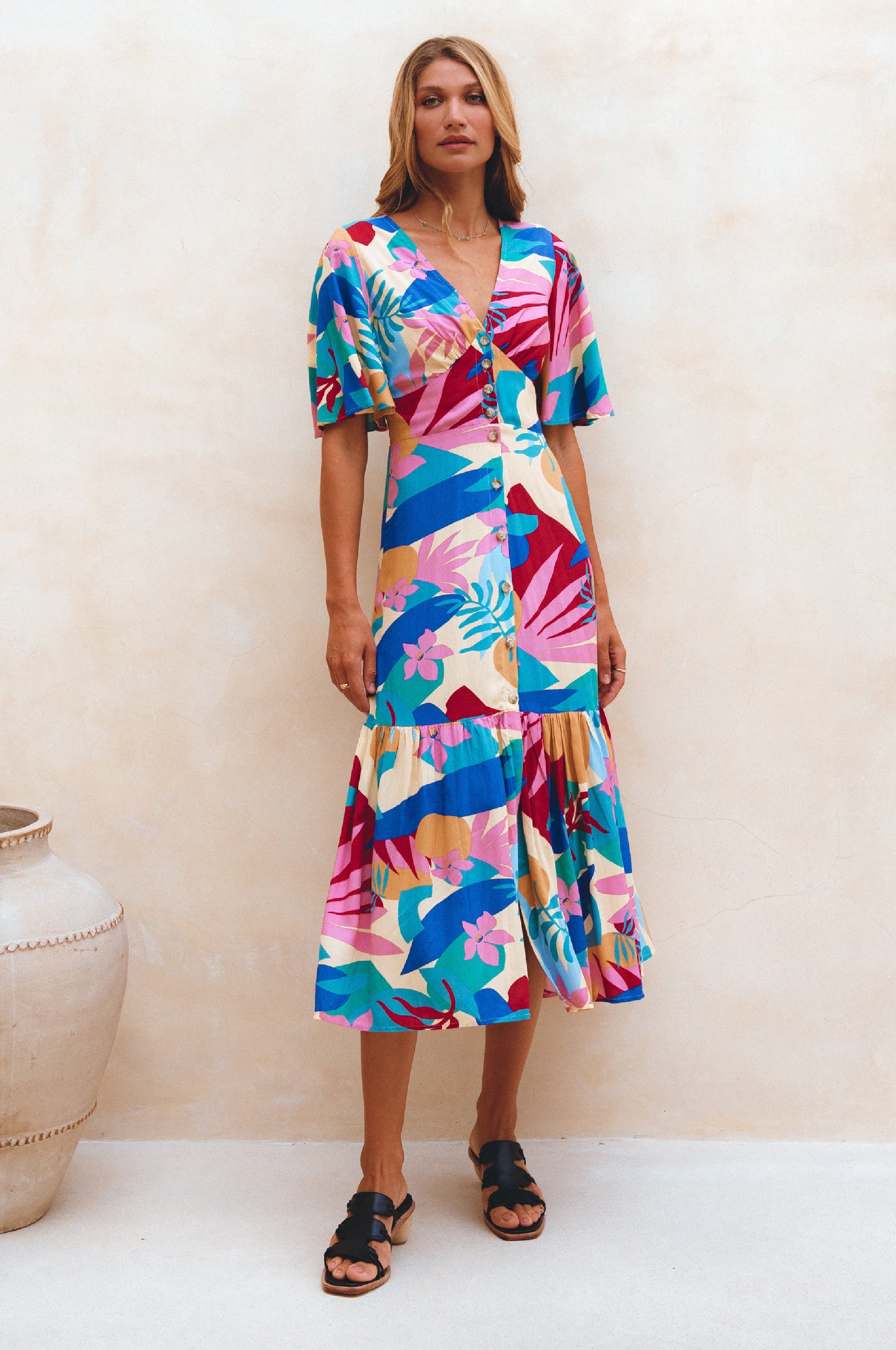 Woman wearing blue, pink and yellow patterned midi dress in a button-down silhouette with a smocked back and fitted front, tiered skirt, and butterfly sleeves standing against a neutral background, front view