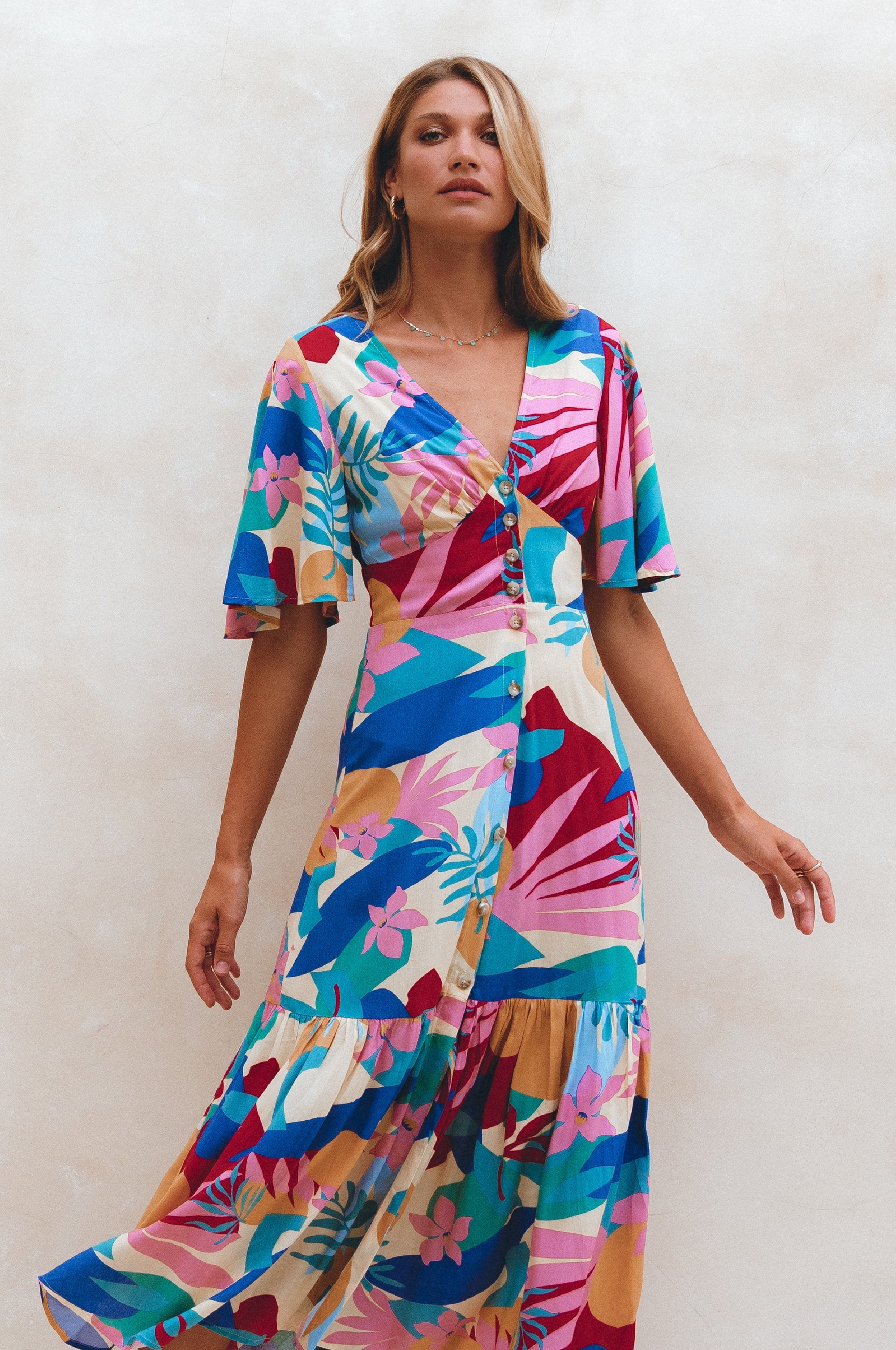 Woman wearing blue, pink and yellow patterned midi dress in a button-down silhouette with a smocked back and fitted front, tiered skirt, and butterfly sleeves standing against a neutral background, front view