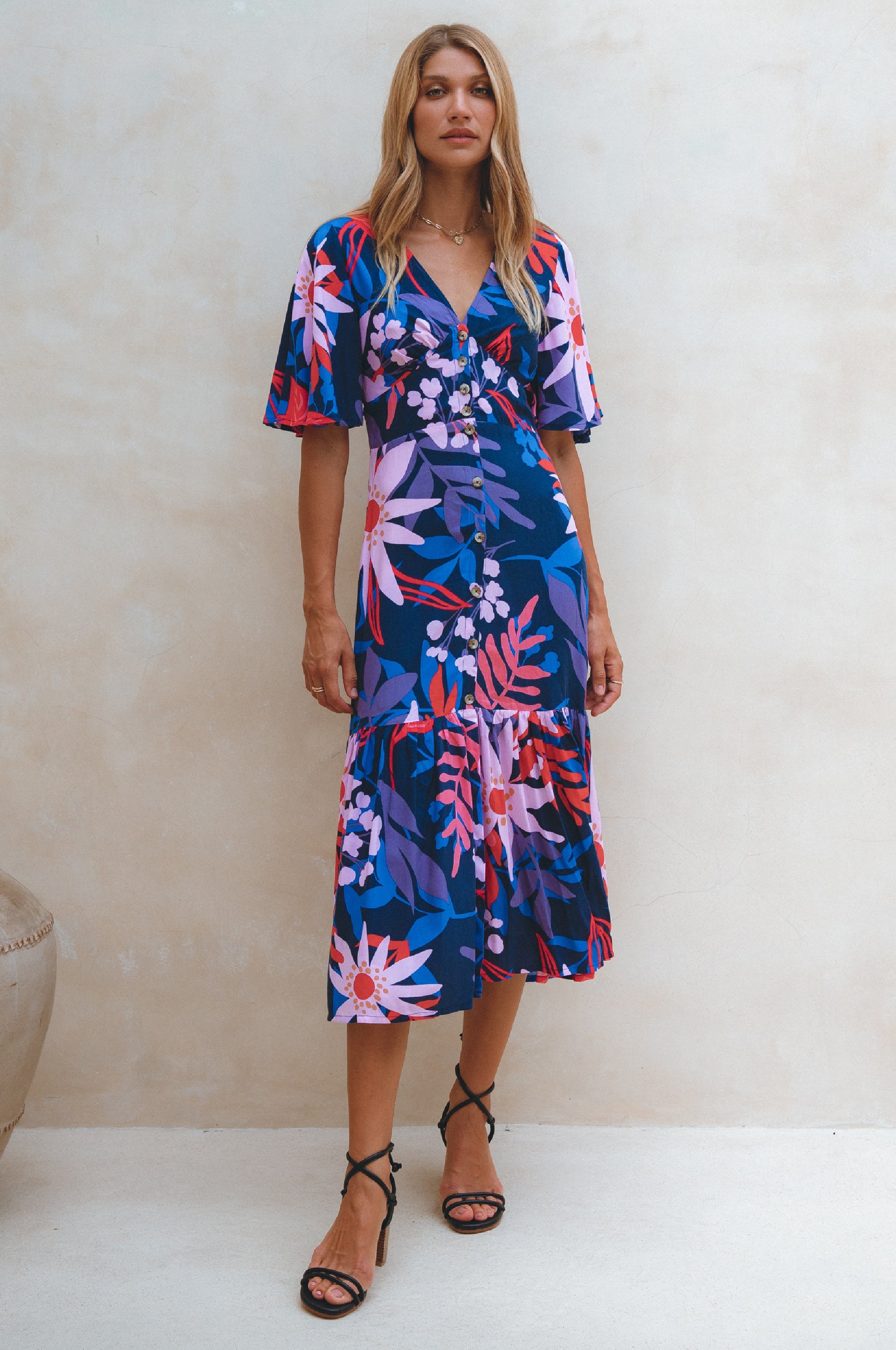 Woman wearing blue, pink patterned midi dress in a button-down silhouette with a smocked back and fitted front, tiered skirt, and butterfly sleeves standing against a neutral background, front view