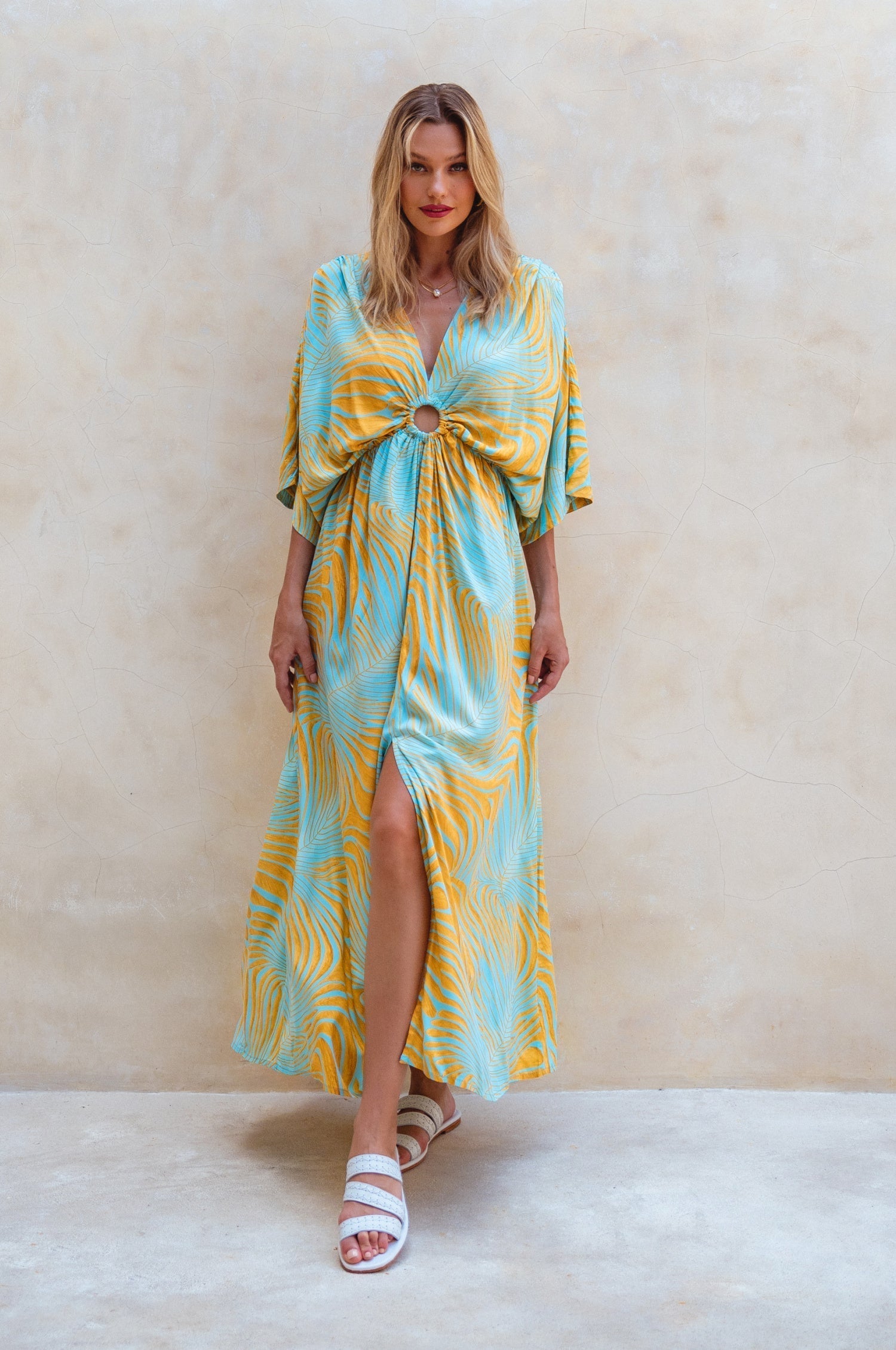 Woman wearing a yellow and blue patterned maxi dress against a beige wall, front view