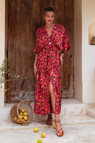 Woman in a vibrant floral maxi dress with v-neckline standing in front of a wooden door with a basket of fruit on the ground, front view
