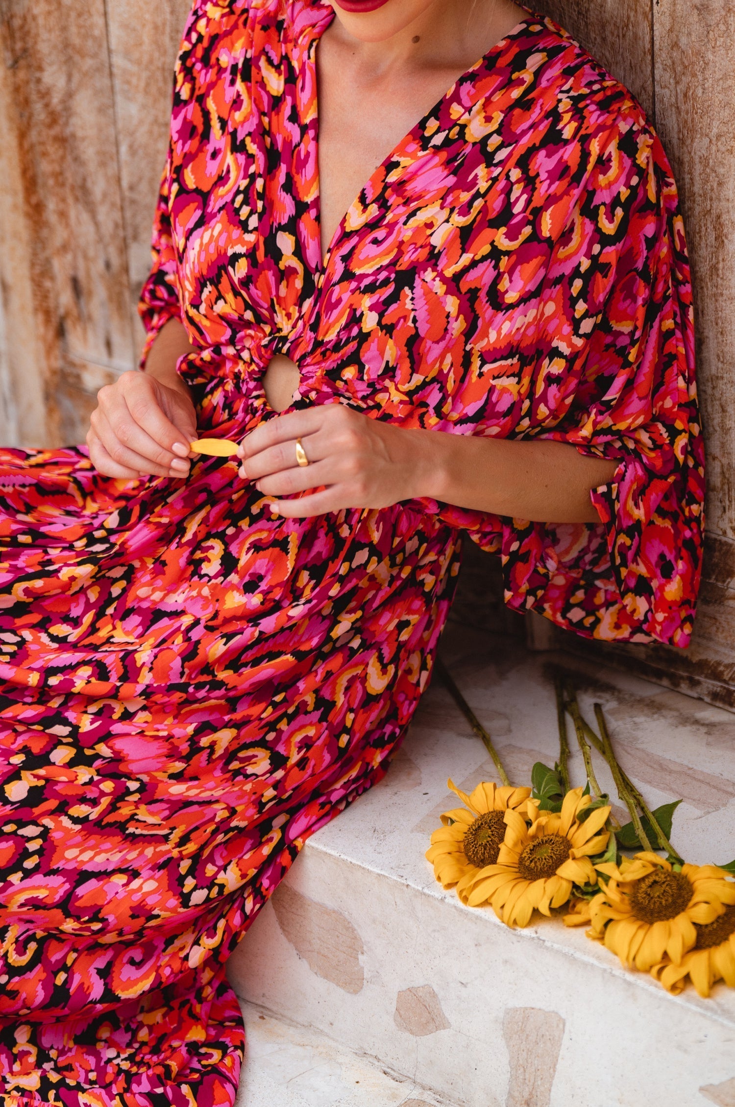 Person wearing a vibrant pink and orange patterned dress sitting on steps with sunflowers.