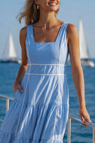Woman in a blue soft cotton tiered midi dress with a square neckline, paneled bodice framed with delicate white trim, and a smocked back, on a boat, front close-up view