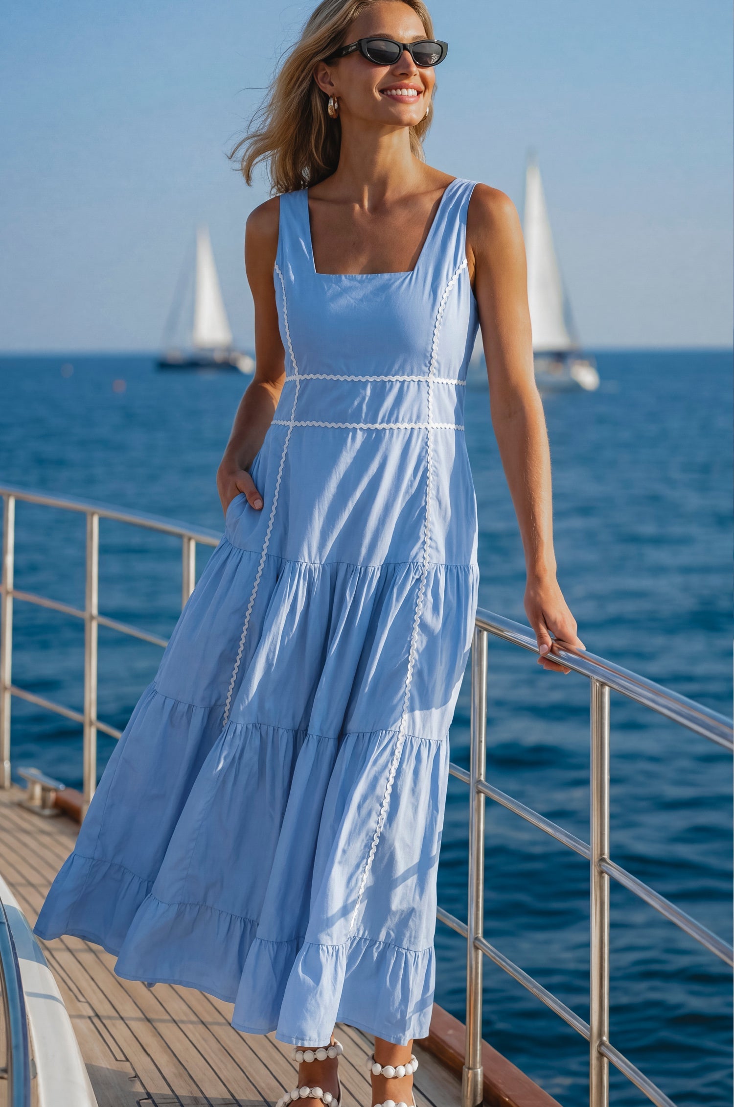 Woman in a blue soft cotton tiered midi dress with a square neckline, paneled bodice framed with delicate white trim, and a smocked back, on a boat, front view