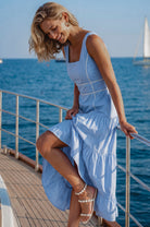 Woman in a blue soft cotton tiered midi dress with a square neckline, paneled bodice framed with delicate white trim, and a smocked back, on a boat, side view