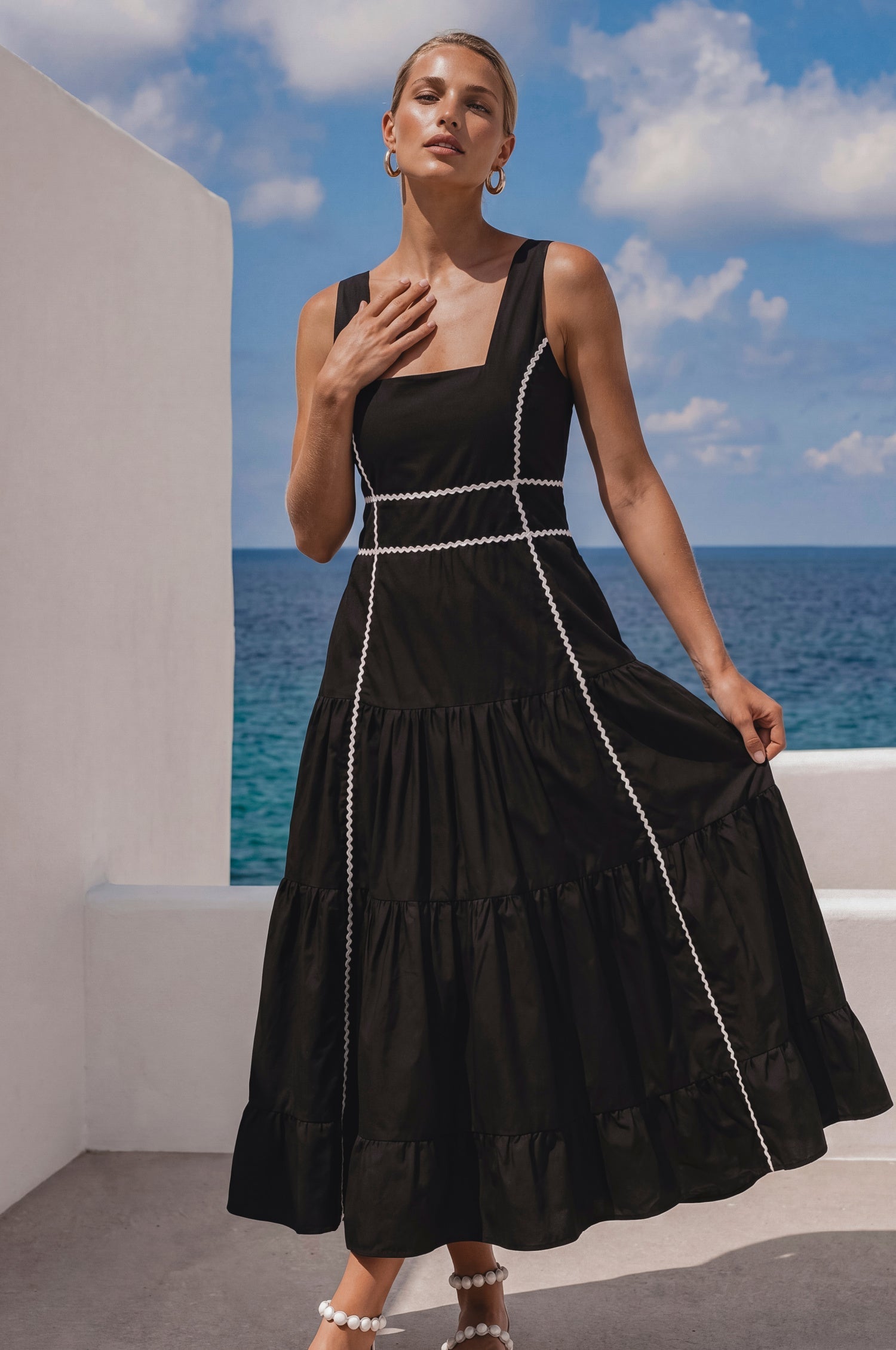 Woman in a black soft cotton tiered midi dress with a square neckline, paneled bodice framed with delicate white trim, and a smocked back, on a boat, front view