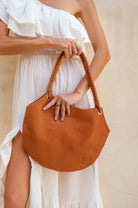 Camel leather handbag held by a person wearing a white dress against a neutral background