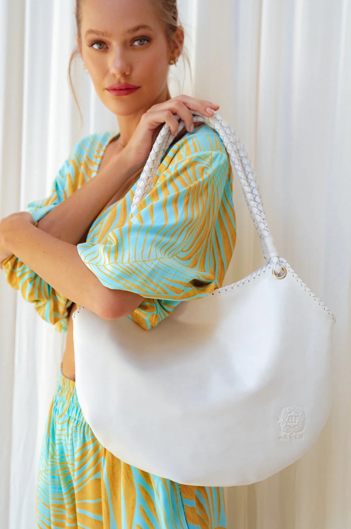 A woman wearing a yellow and turquoise patterned dress holds a large white leather shoulder bag with a braided strap against a white curtain background.