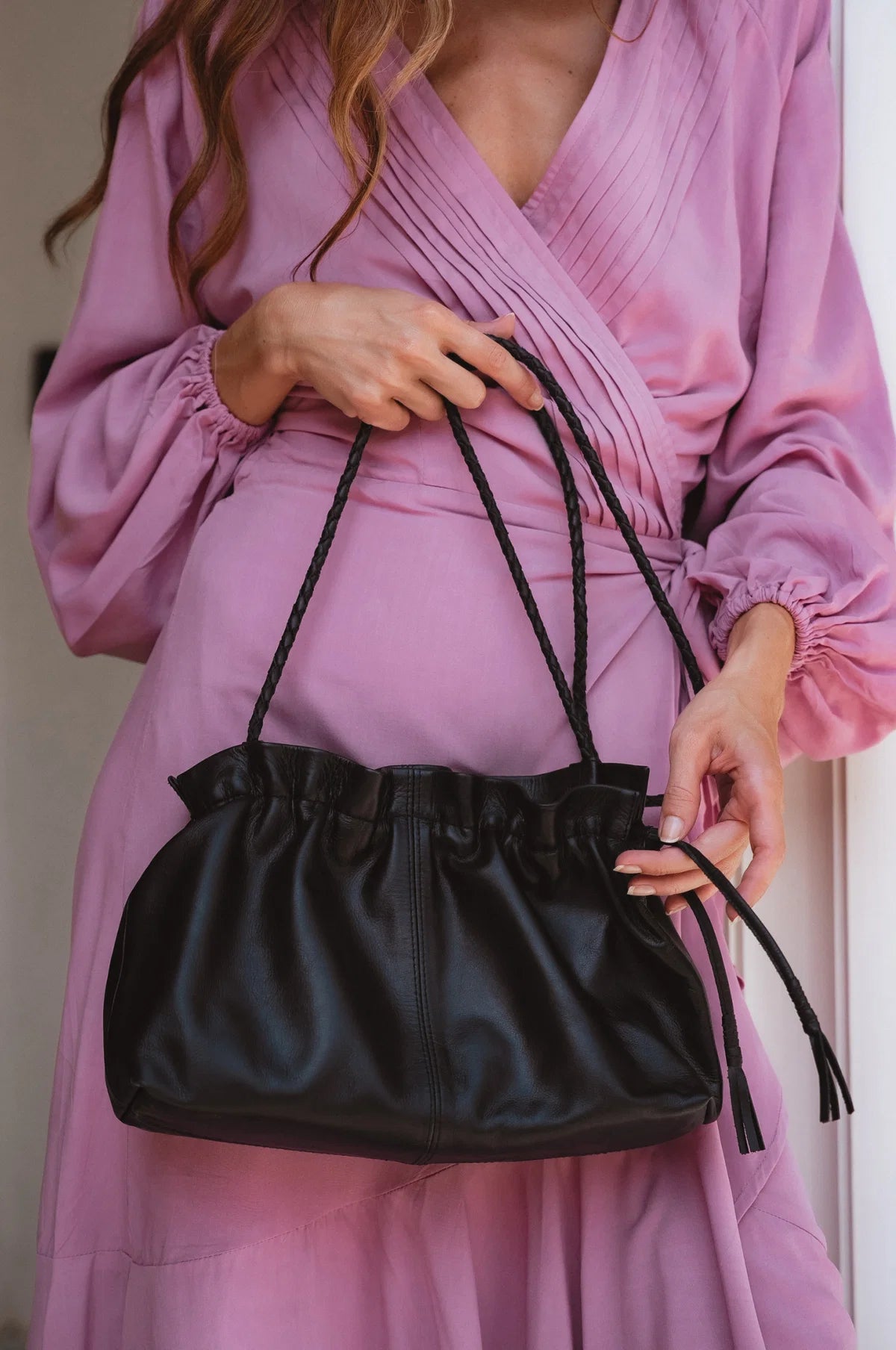 Person holding a black leather handbag with leather rope strap wearing a pink dress.