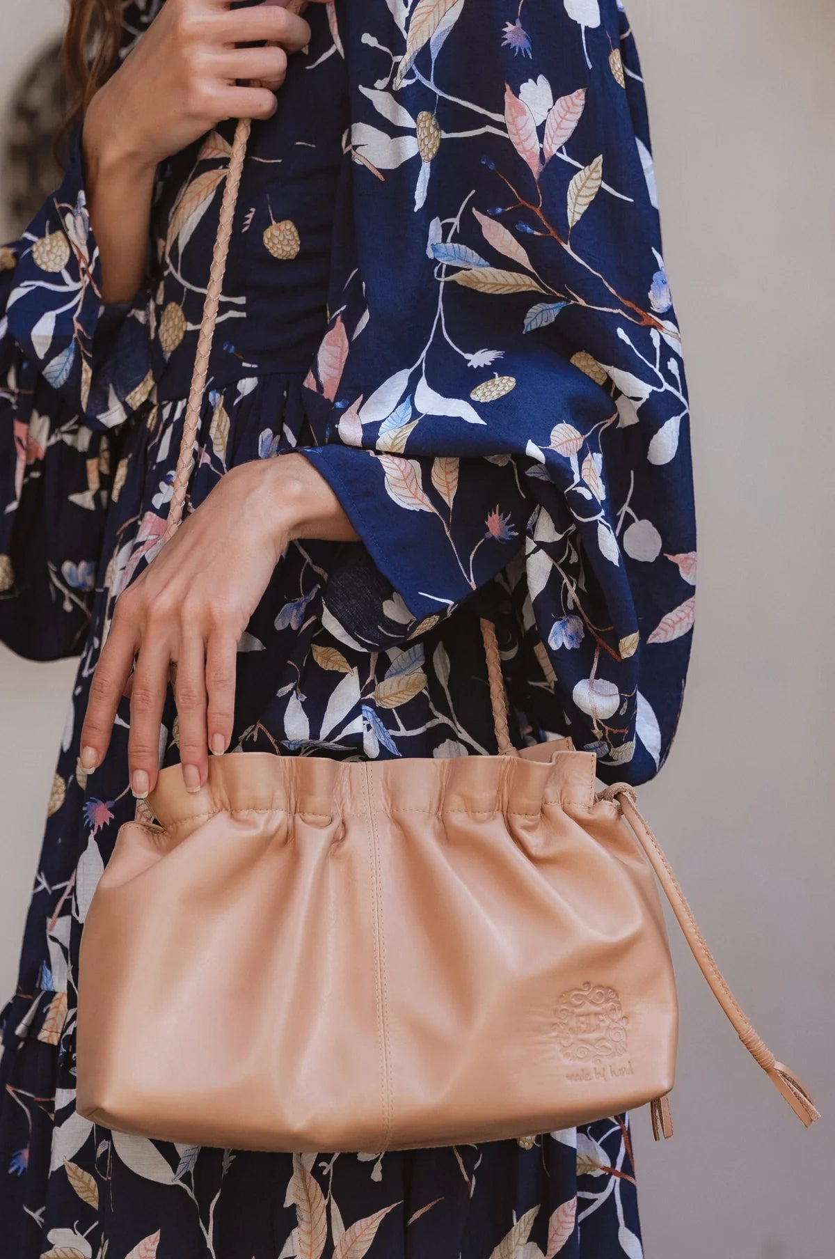 Person holding a beige leather crossbody handbag with drawstring top and braided strap with a floral-patterned garment in the background