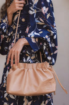 Person holding a beige leather crossbody handbag with drawstring top and braided strap with a floral-patterned garment in the background