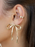 Close-up of woman wearing gold-tone bow earring with wavy ribbon tails.