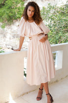Woman in a cream midi dress with \voluminous skirt, puff sleeves, and cutouts at the waist framed by wraparound ties standing on a balcony with greenery in the background