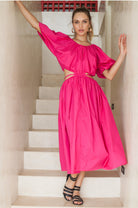 Woman in a bright pink midi dress with \voluminous skirt, puff sleeves, and cutouts at the waist framed by wraparound ties standing on a staircase