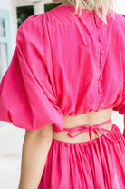 Woman in a bright pink midi dress with \voluminous skirt, puff sleeves, and cutouts at the waist framed by wraparound ties, back view.