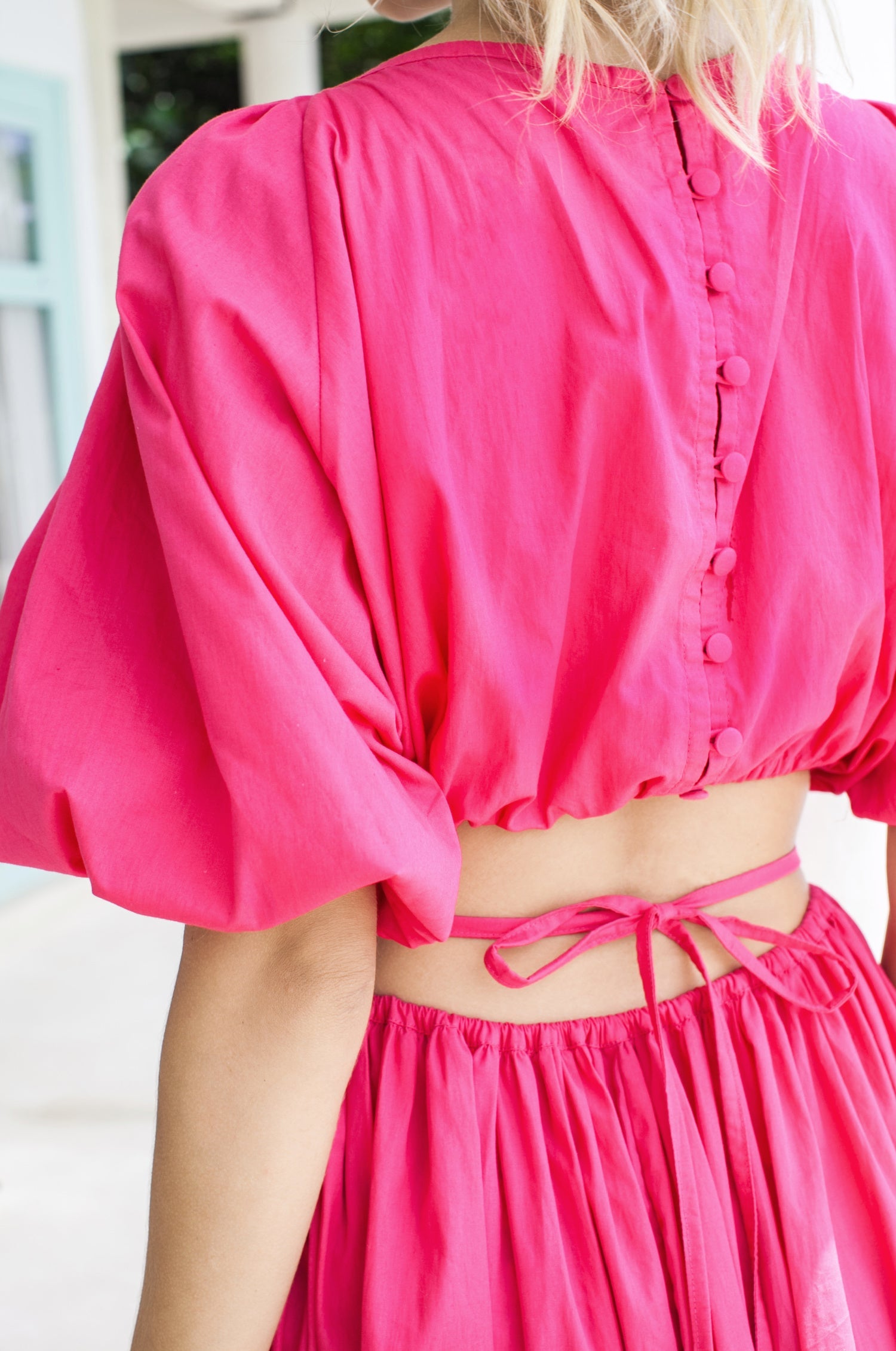 Woman in a bright pink midi dress with \voluminous skirt, puff sleeves, and cutouts at the waist framed by wraparound ties, back view.