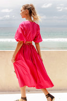 Woman in a bright pink midi dress with \voluminous skirt, puff sleeves, and cutouts at the waist framed by wraparound ties standing on a beach with ocean view, back view.