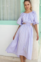 Woman wearing a light purple dress with cut-out details, standing against a light-colored wall.