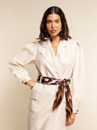 Woman wearing a white coat with a patterned silk scarf as a belt against a beige background