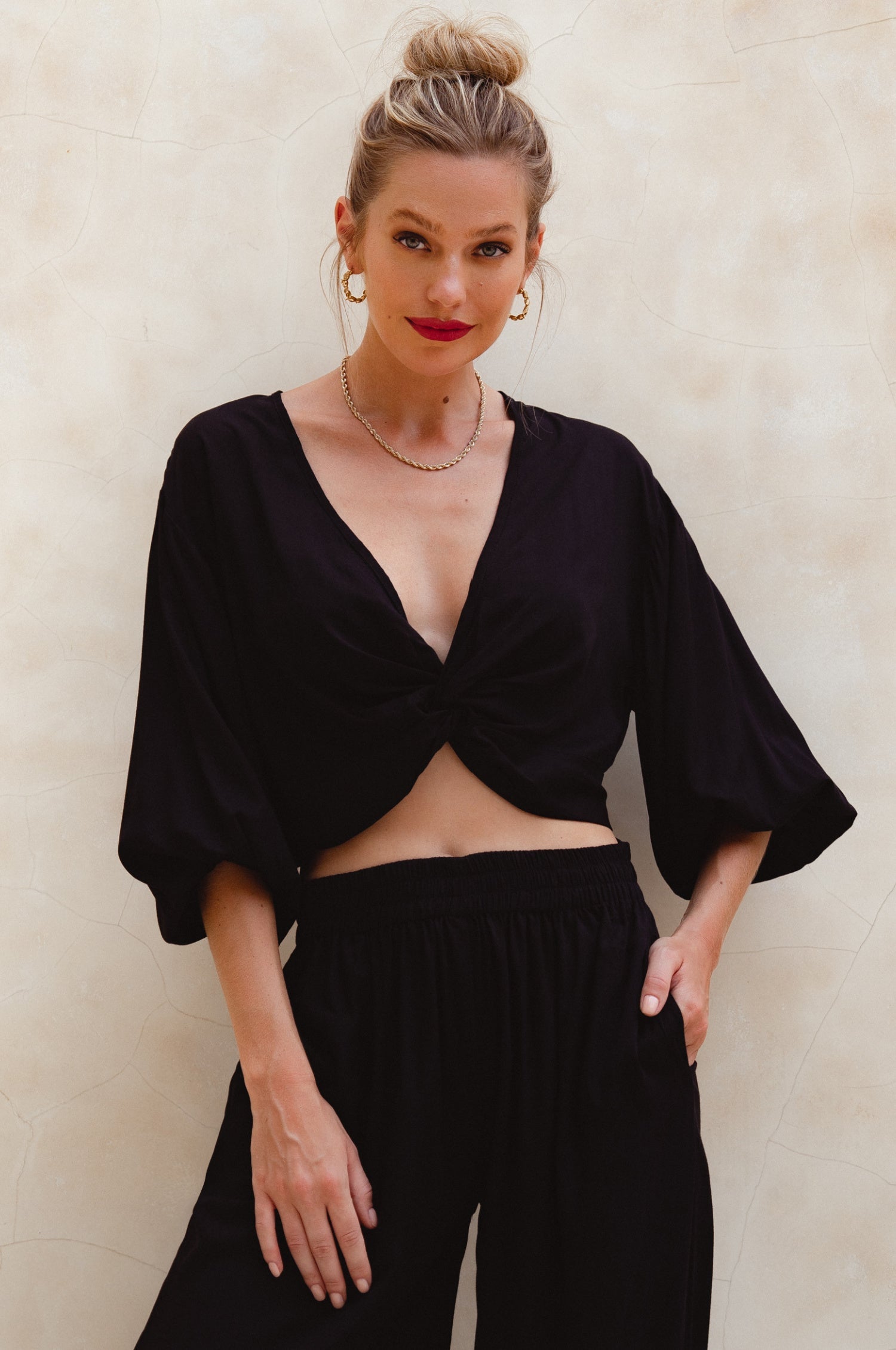 Woman wearing a black unlined slip-on cropped style top with twisted detailing on the front and elasticated sleeves and matching pants against a plain background