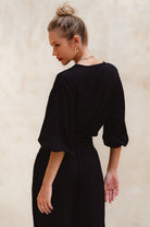 Woman wearing a black unlined slip-on cropped style top with twisted detailing on the front and elasticated sleeves and matching pants dress against a beige wall, back view