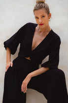 Woman wearing a black unlined slip-on cropped style top with twisted detailing on the front and elasticated sleeves and matching pants against a neutral background