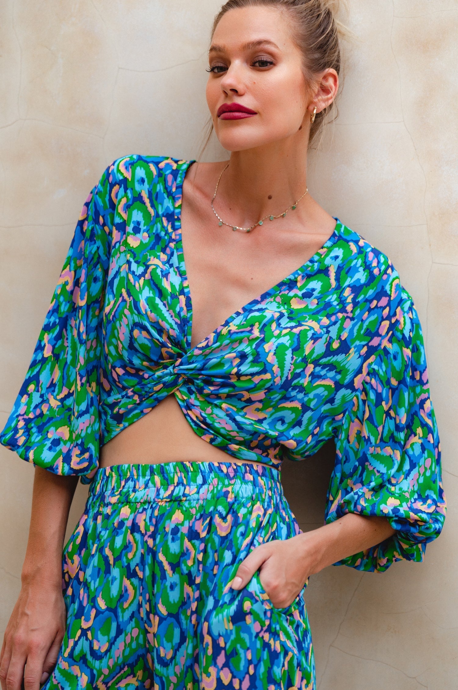 Woman wearing a colorful blue, pink and green slip-on cropped style top with twisted detailing on the front and elasticated sleeves and matching pants against a beige wall, closeup