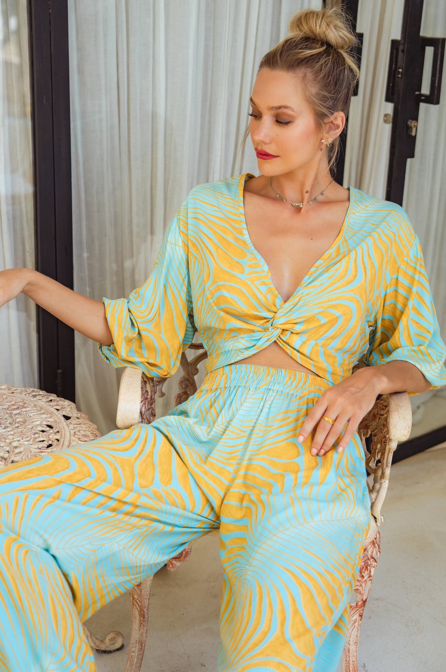 Woman wearing a yellow and blow patterned slip-on cropped style top with twisted detailing on the front and elasticated sleeves and matching pants sitting on a chair.