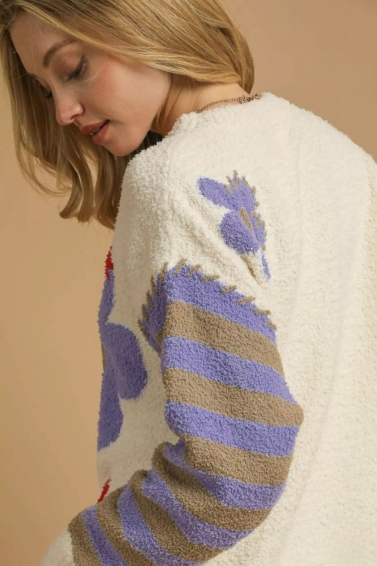 Close-up of cream plush knit sweater showing lavender and taupe striped sleeves and floral detailing near the shoulder.