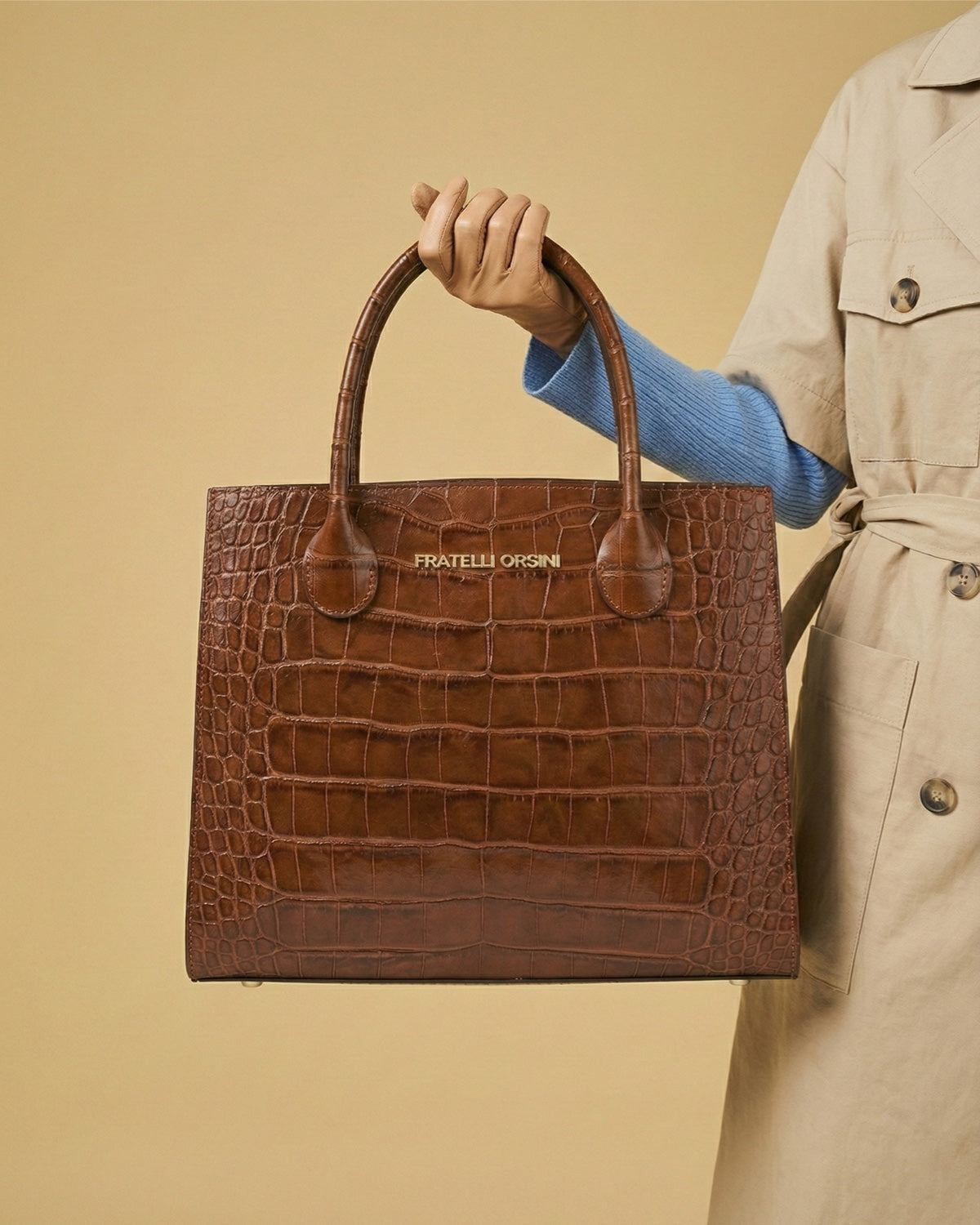 Brown crocodile-patterned handbag held by a person wearing a beige trench coat on a beige background
