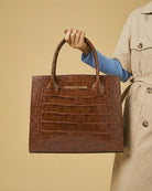 Brown crocodile-patterned handbag held by a person wearing a beige trench coat on a beige background
