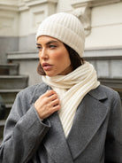 Woman wearing an ivory knit beanie and scarf with a gray coat outdoors.
