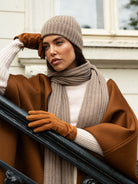 Woman wearing a brown coat, beige hat, gloves, and scarf standing outdoors.