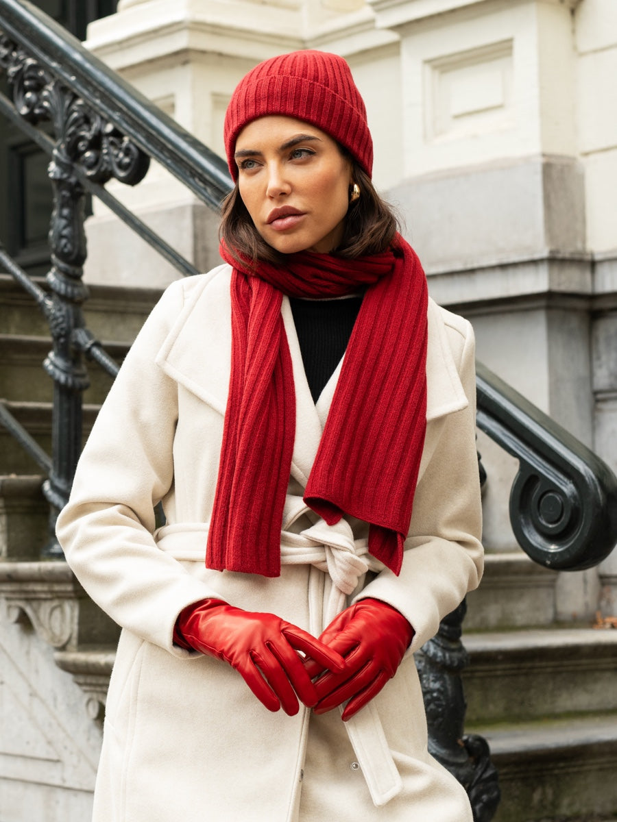 Woman wearing a red cashmere beanie, scarf, and gloves with a white coat against a stone building background.