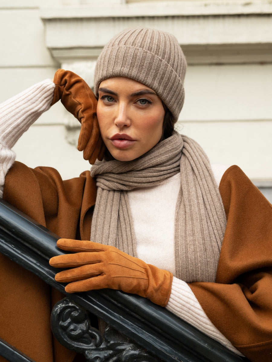 Woman wearing a brown coat, beige beanie, scarf, and gloves, standing next to a car.