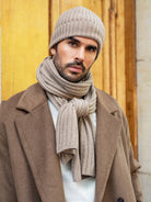 Man wearing a beige knit hat and scarf with a brown coat against a wooden background