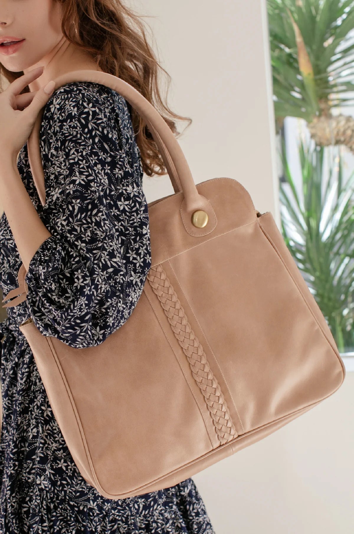 A woman wearing a black and white floral dress carries a large beige leather tote bag over her shoulder. The bag features a structured rectangular shape, a top handle, a gold snap button closure, and a braided decorative detail running vertically on the front.
