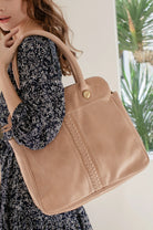 A woman wearing a black and white floral dress carries a large beige leather tote bag over her shoulder. The bag features a structured rectangular shape, a top handle, a gold snap button closure, and a braided decorative detail running vertically on the front.
